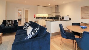 A modern living room with a blue sofa set, a dining table with chairs, a kitchen area with white cabinets and a microwave, and a ceiling light fixture.