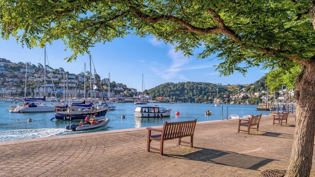 A waterfront promenade with benches, boats, and a view of a hillside town.