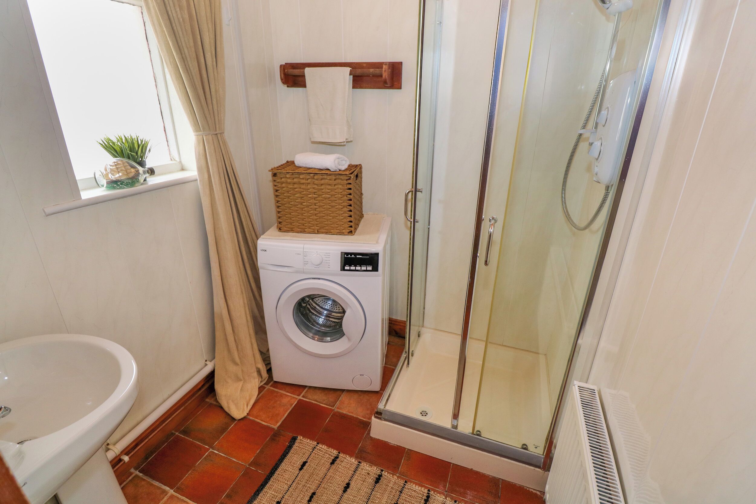 Un baño pequeño con lavadora, mampara de vidrio, lavamanos y una ventana con una planta.