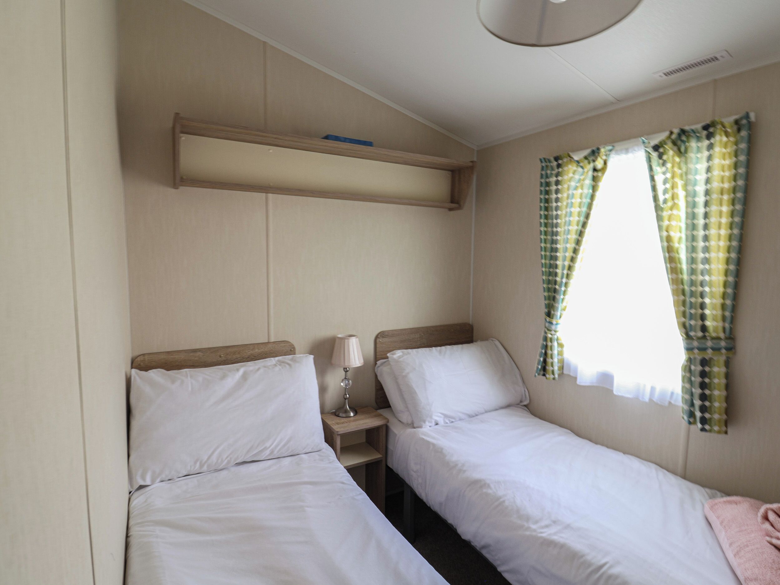 A small, clean room with two single beds, a bedside table with a lamp, a shelf above the beds, and a window with patterned curtains.