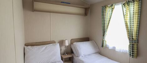A small, clean room with two single beds, a bedside table with a lamp, a shelf above the beds, and a window with patterned curtains.