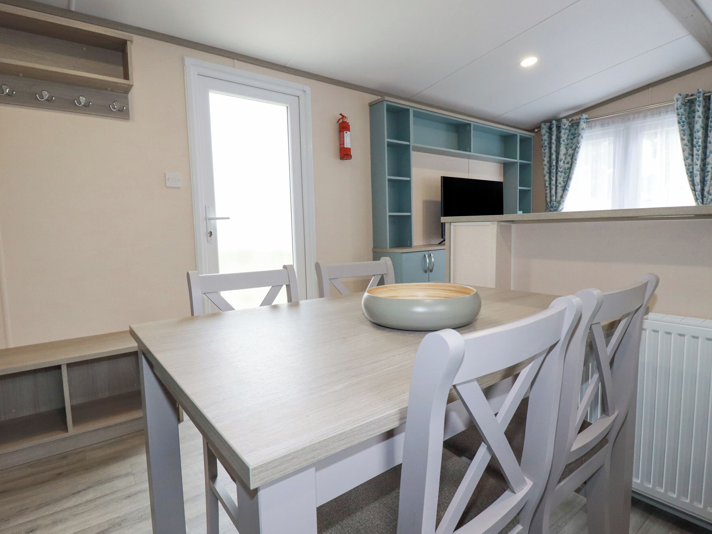 A dining area with a wooden table, white chairs, a bowl, a fire extinguisher, and a kitchen area with blue cabinets and a window with curtains.