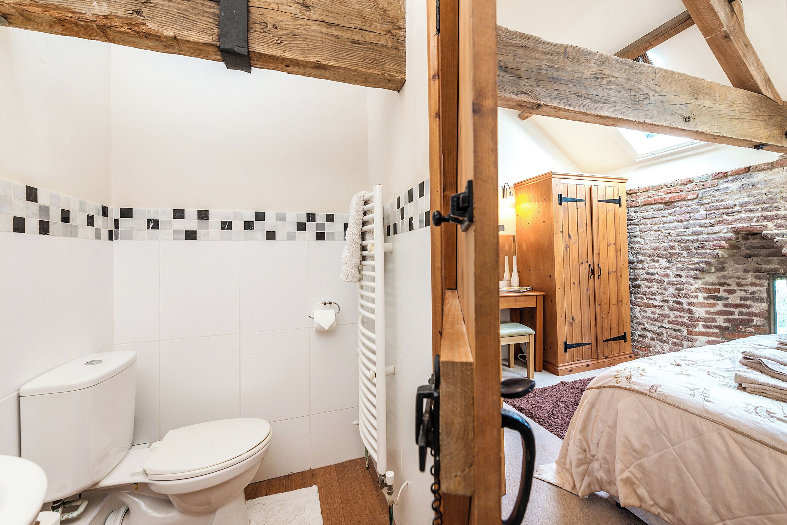 Une salle de bain avec des toilettes blanches, un porte-serviettes et une porte en bois donnant sur une chambre à coucher équipée d’un lit et d’une armoire en bois.