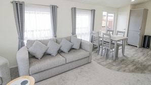 A living room with a grey sofa, a dining table with chairs, and a kitchen area with a fridge. - Long Acres Lodge (Evesham)