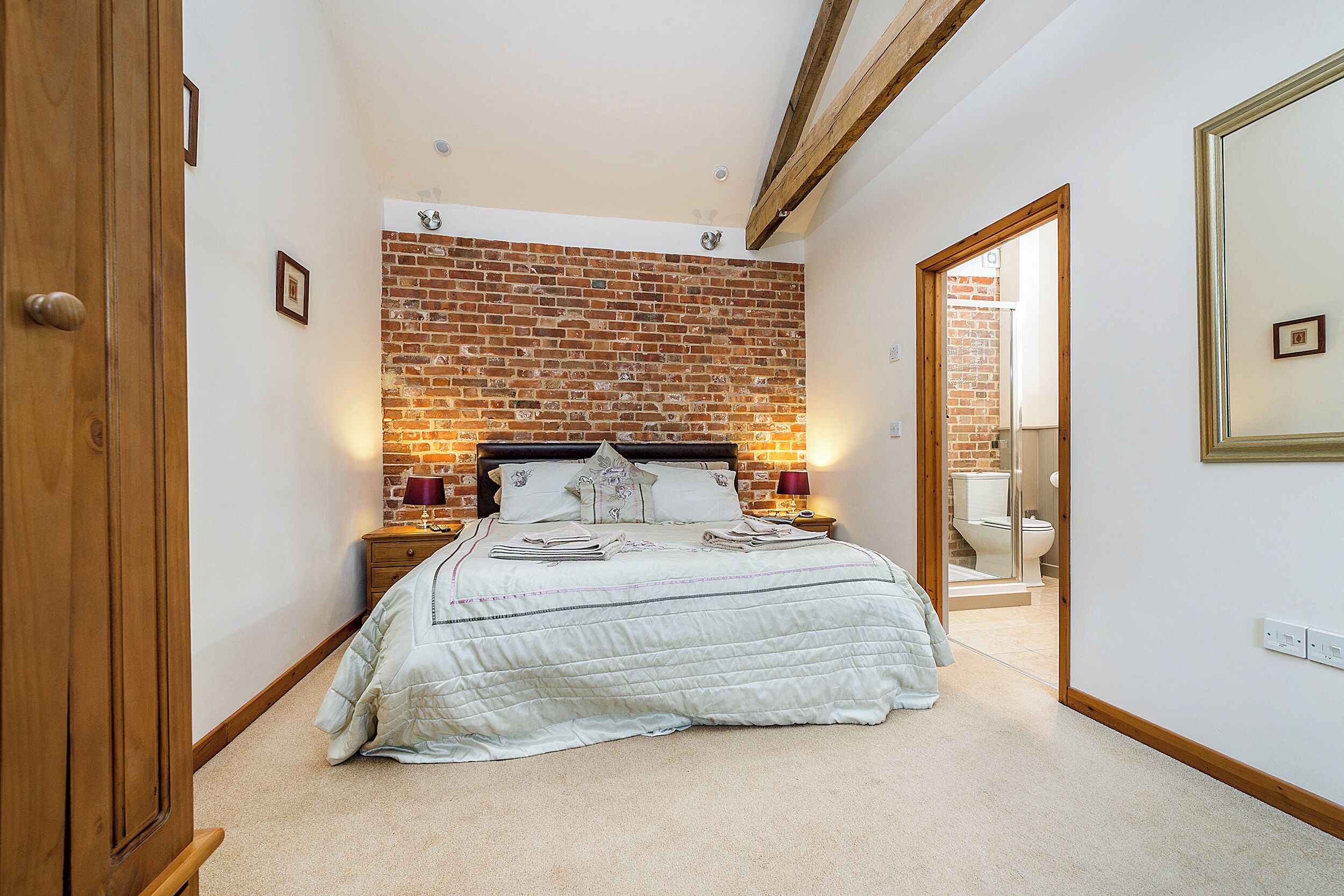 Een slaapkamer met een bakstenen muur, een bed met sprei, nachtkastjes en een spiegel.