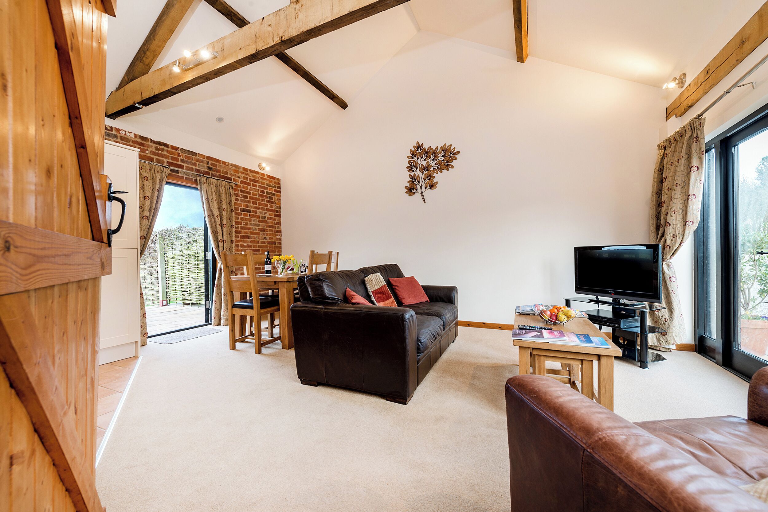 A spacious living room with a black leather sofa, a wooden coffee table, and a television. A brick wall and wooden beams are visible, and there is a large window with curtains.