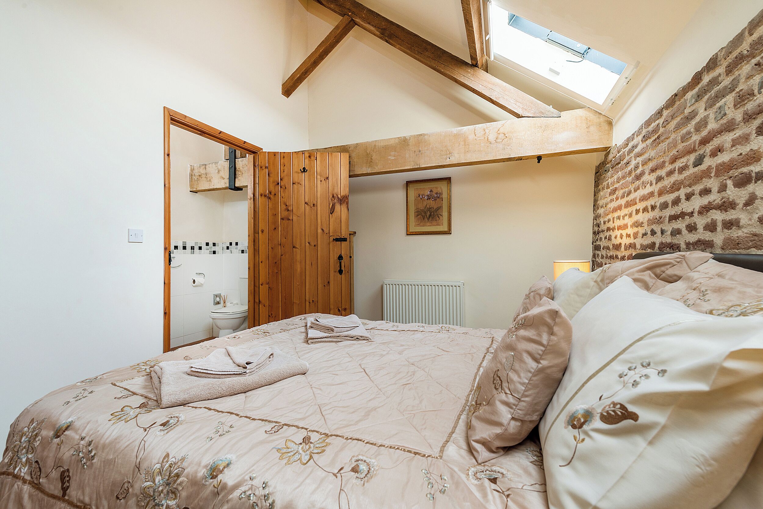 Une chambre avec un plafond en pente, un puits de lumière, un lit avec une literie à motifs floraux, un radiateur et une porte en bois.