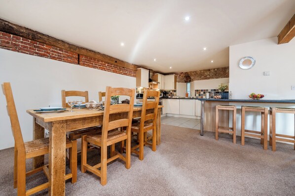 A dining area with a wooden table and chairs, a kitchen in the background, and a brick wall. - Teal (Beccles)