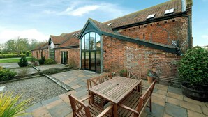 A brick building with a large patio area, a wooden table and chairs, and a glass door entrance. - Teal (Beccles)