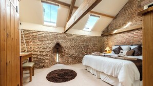 A bedroom with a stone wall, wooden beams, a bed with white bedding, a small round rug, and a wooden nightstand with a lamp. - Teal (Beccles)
