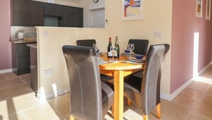 A dining area with a table set for four, a kitchen in the background, a clock on the wall, and a poster on the wall. - The Lodge at Redlands (Lyme Regis)
