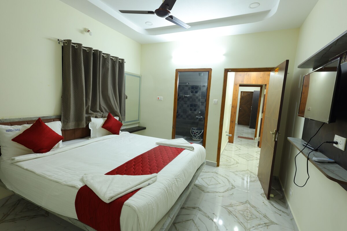 Habitación de hotel con una cama grande, un ventilador de techo, un televisor y un baño visible a través de una puerta abierta.