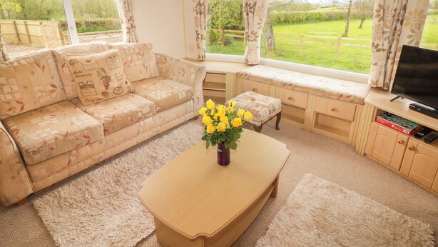 A living room with a sofa, a coffee table with a vase of yellow flowers, a television, and a view of a green landscape through large windows. - Grandads Shack (Pontypool)