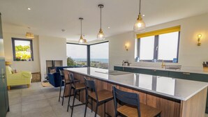 Une cuisine moderne dotée d’un grand îlot central, de tabourets de bar, de luminaires suspendus et offrant une vue sur la mer.
