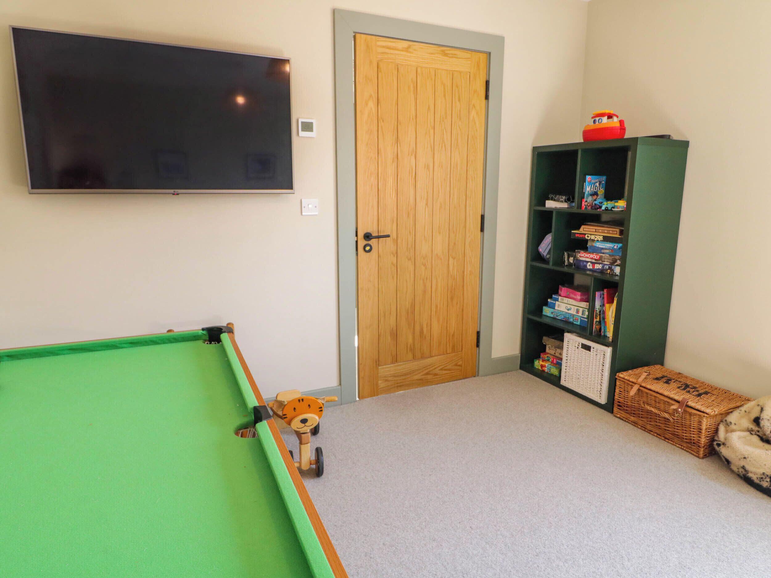 A room with a pool table, a television mounted on the wall, a wooden door, and a bookshelf with board games.