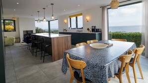 Une cuisine moderne dotée d’un îlot central, d’un coin repas avec une grande table et des chaises, et offrant une vue sur la mer.