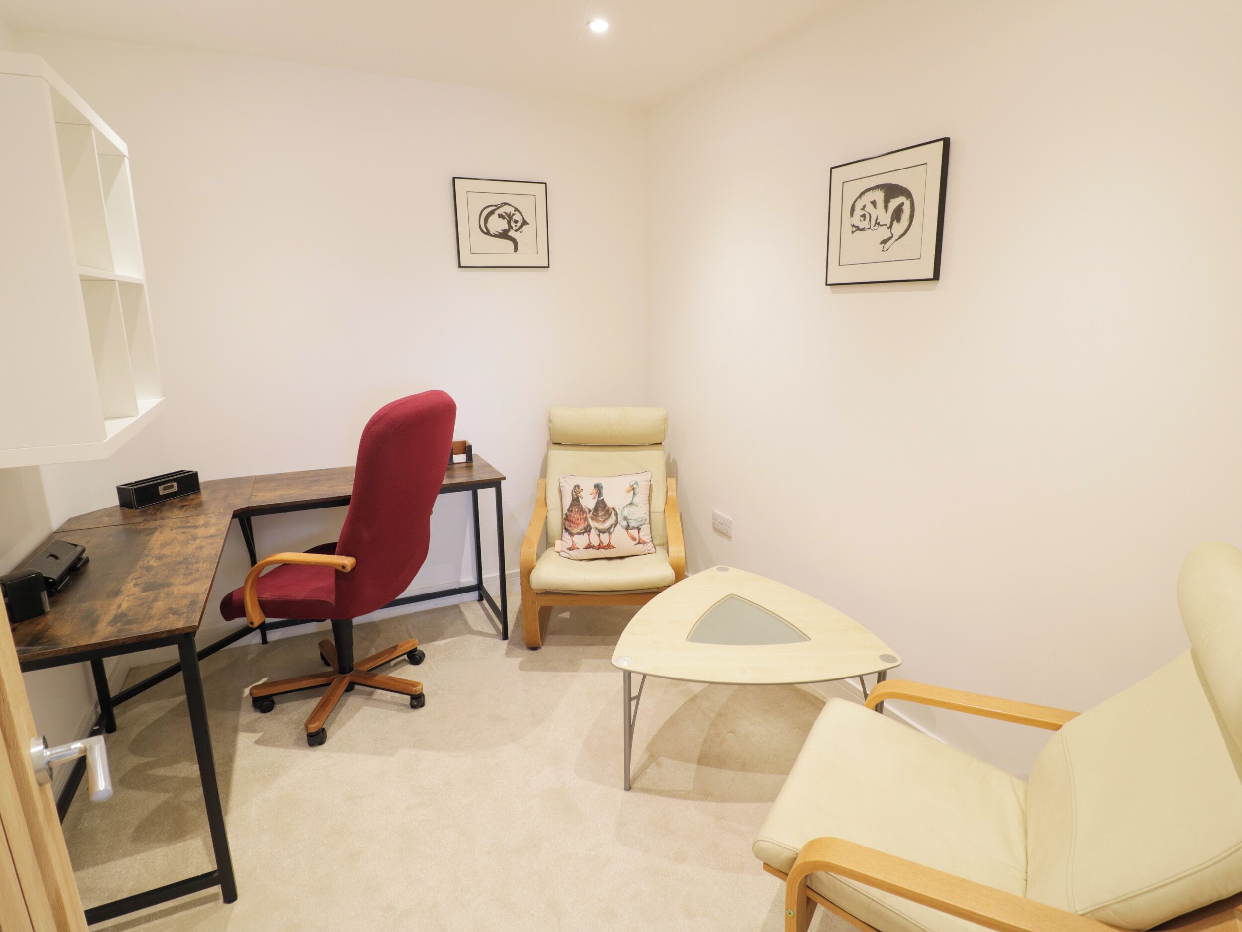 A small, well-lit room with a desk, two chairs, and two framed pictures on the wall.
