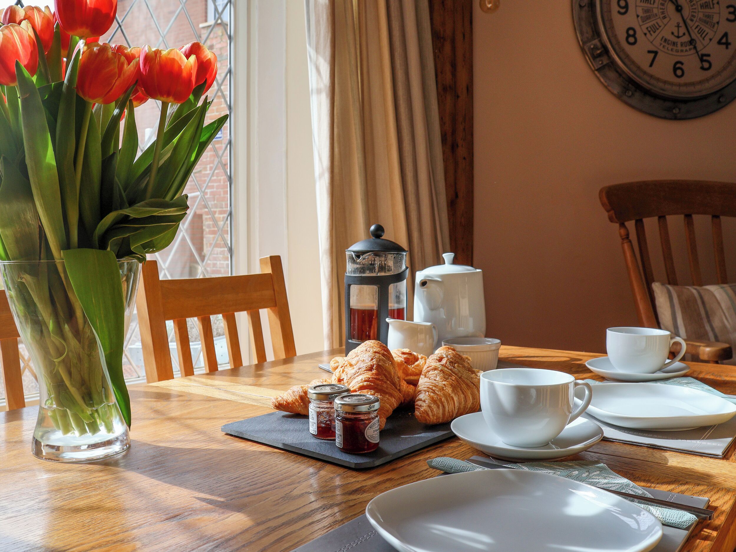 A wooden dining table with croissants, a teapot, and a cup, set against a window with a view of a brick wall and a clock.