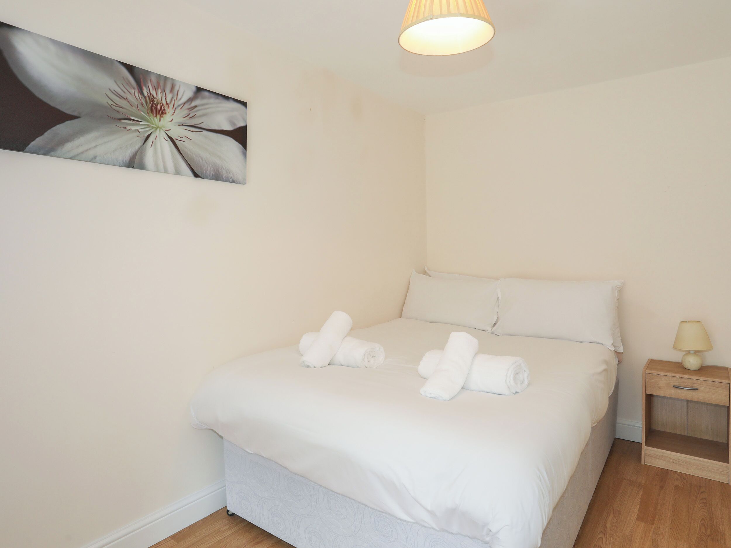 A neatly made bed with white linens and rolled towels, a bedside table with a lamp, and a wall-mounted painting of a flower.