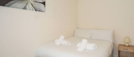 A neatly made bed with white linens and rolled towels, a bedside table with a lamp, and a wall-mounted painting of a flower.