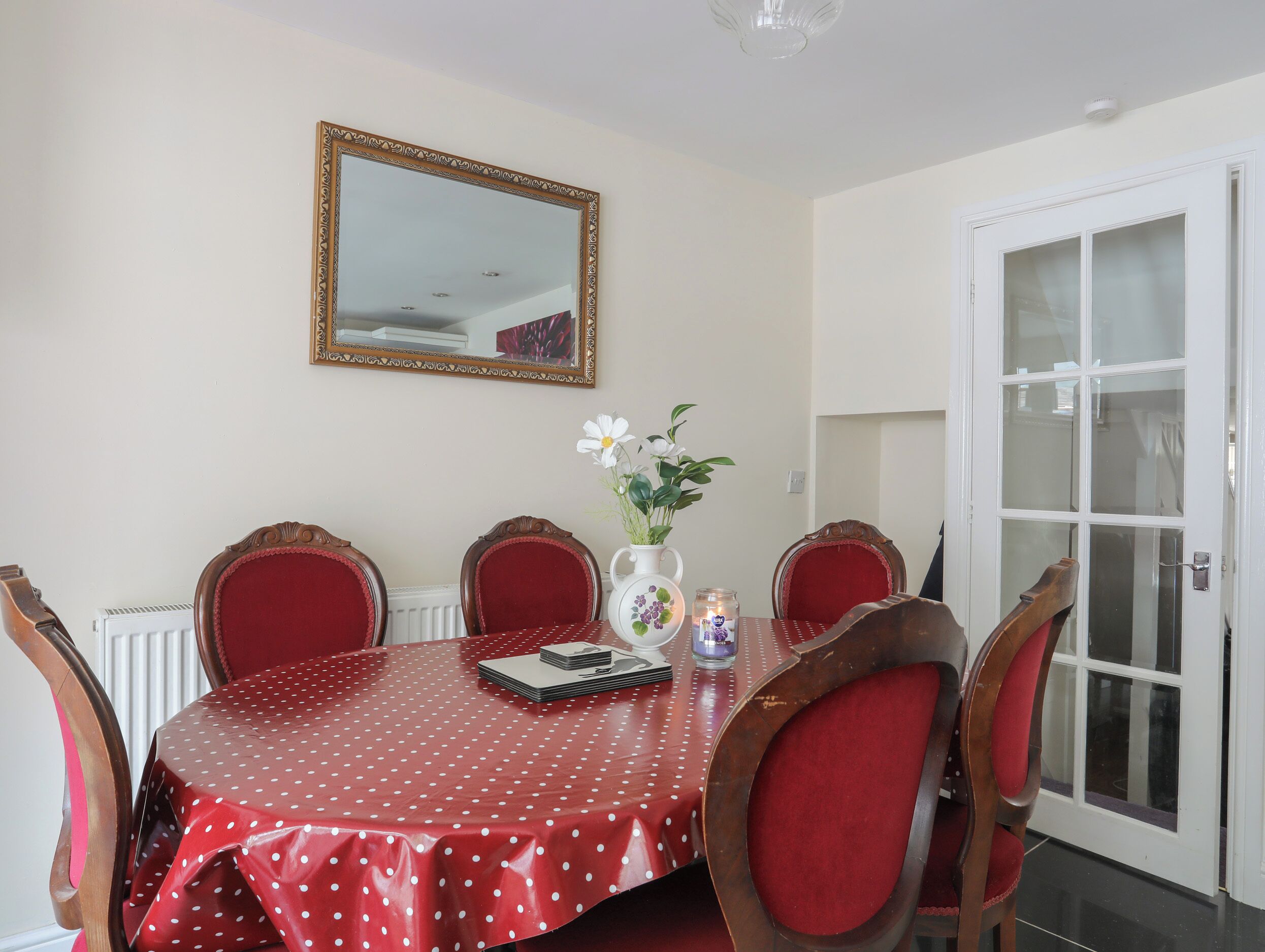 A dining area with a red table covered in white polka dots, surrounded by six wooden chairs with red cushions. A white vase with flowers and a small candle are on the table.