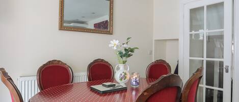 Un comedor con una mesa roja cubierta de lunares blancos, rodeado por seis sillas de madera con cojines rojos. En la mesa hay un florero blanco con flores y una vela pequeña.