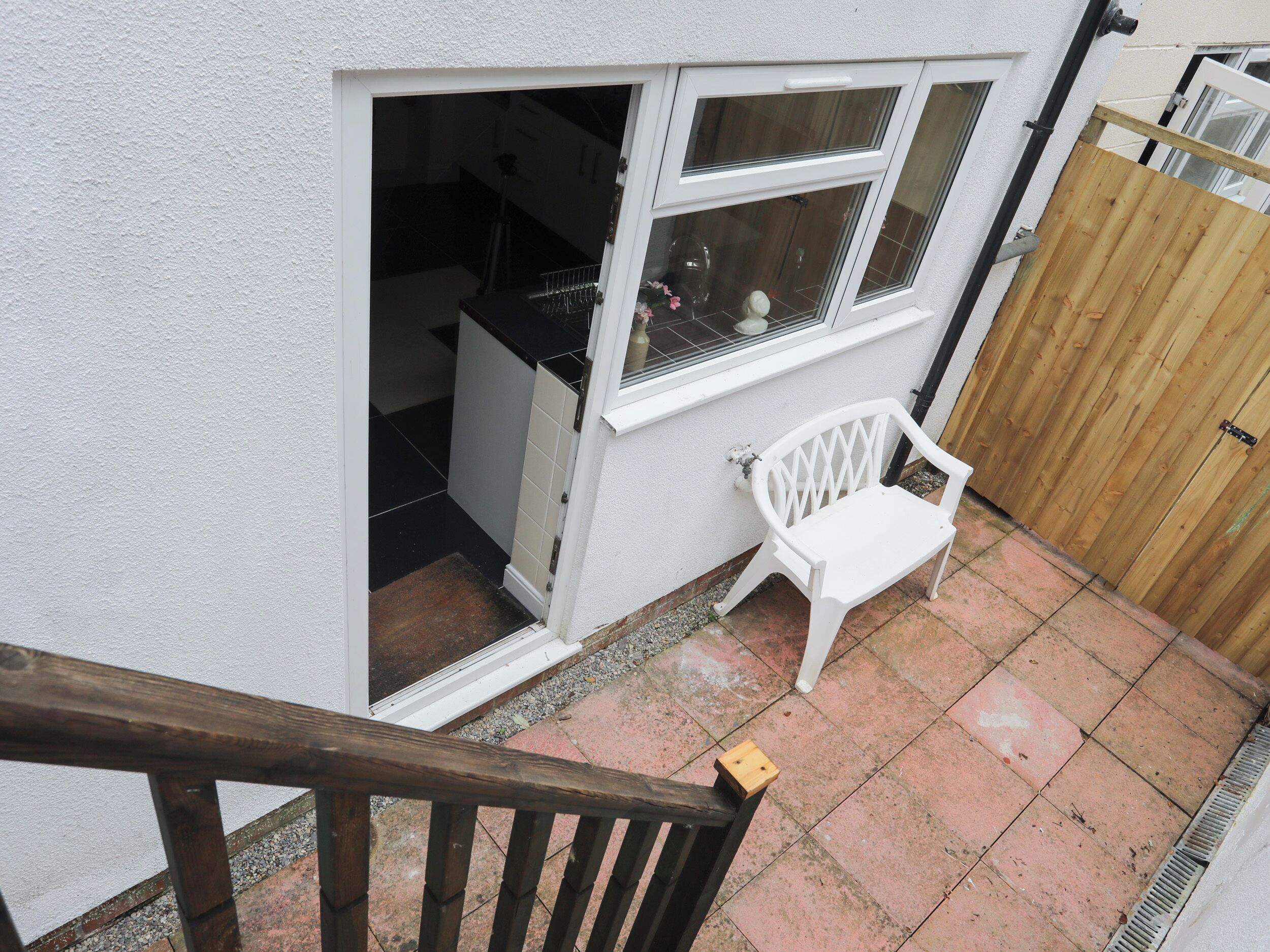 A small patio area with a white plastic chair, a wooden fence, and a white wall with a window.