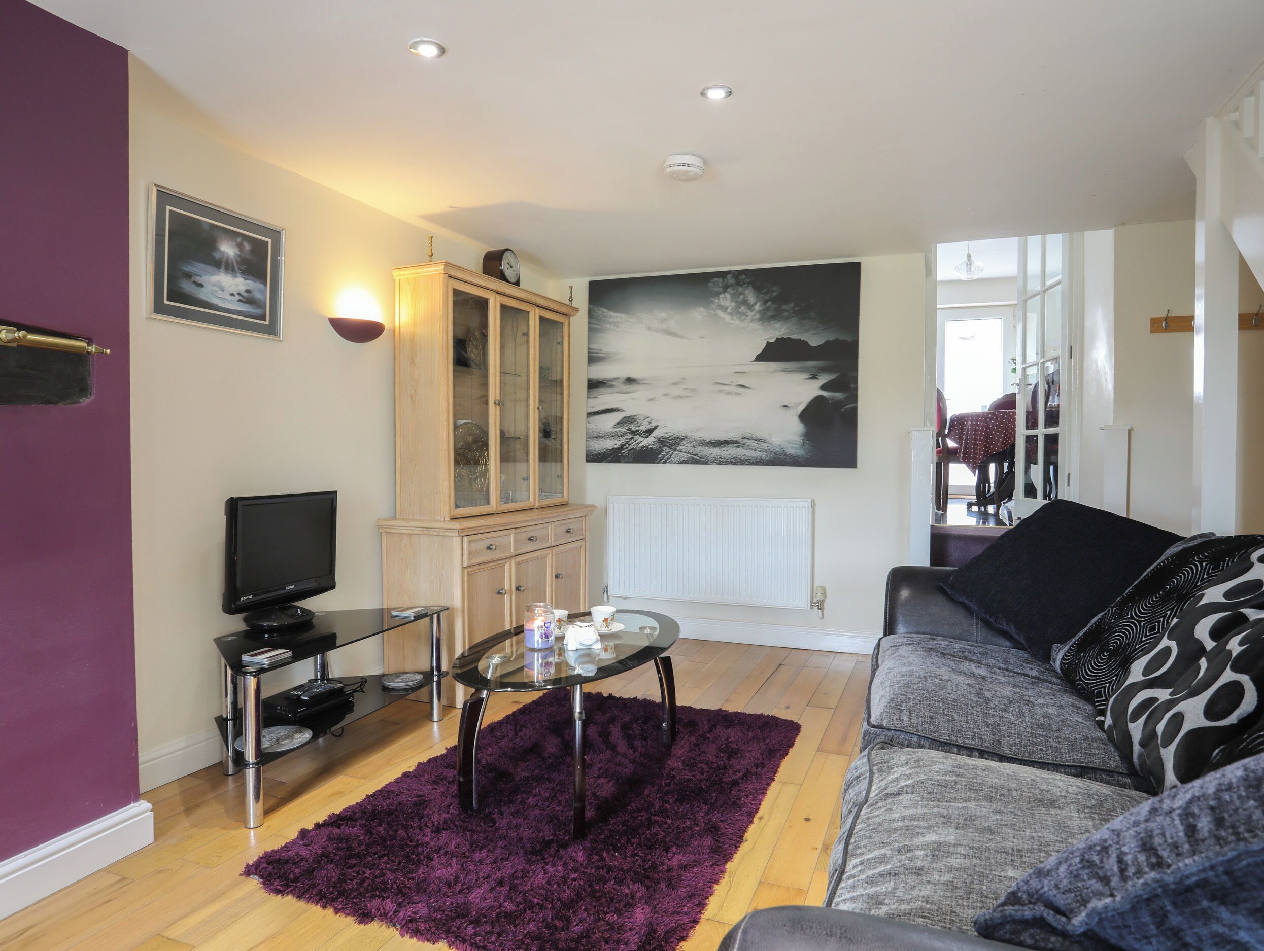 A living room with a sofa, a glass coffee table, a purple rug, a wooden cabinet, a television, and a large painting on the wall.