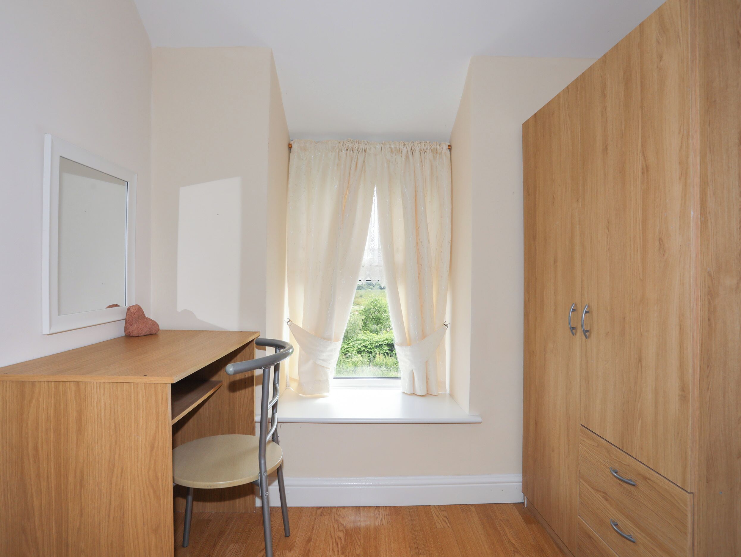 Una habitación pequeña con un escritorio de madera, una silla, un armario y una ventana con cortinas.