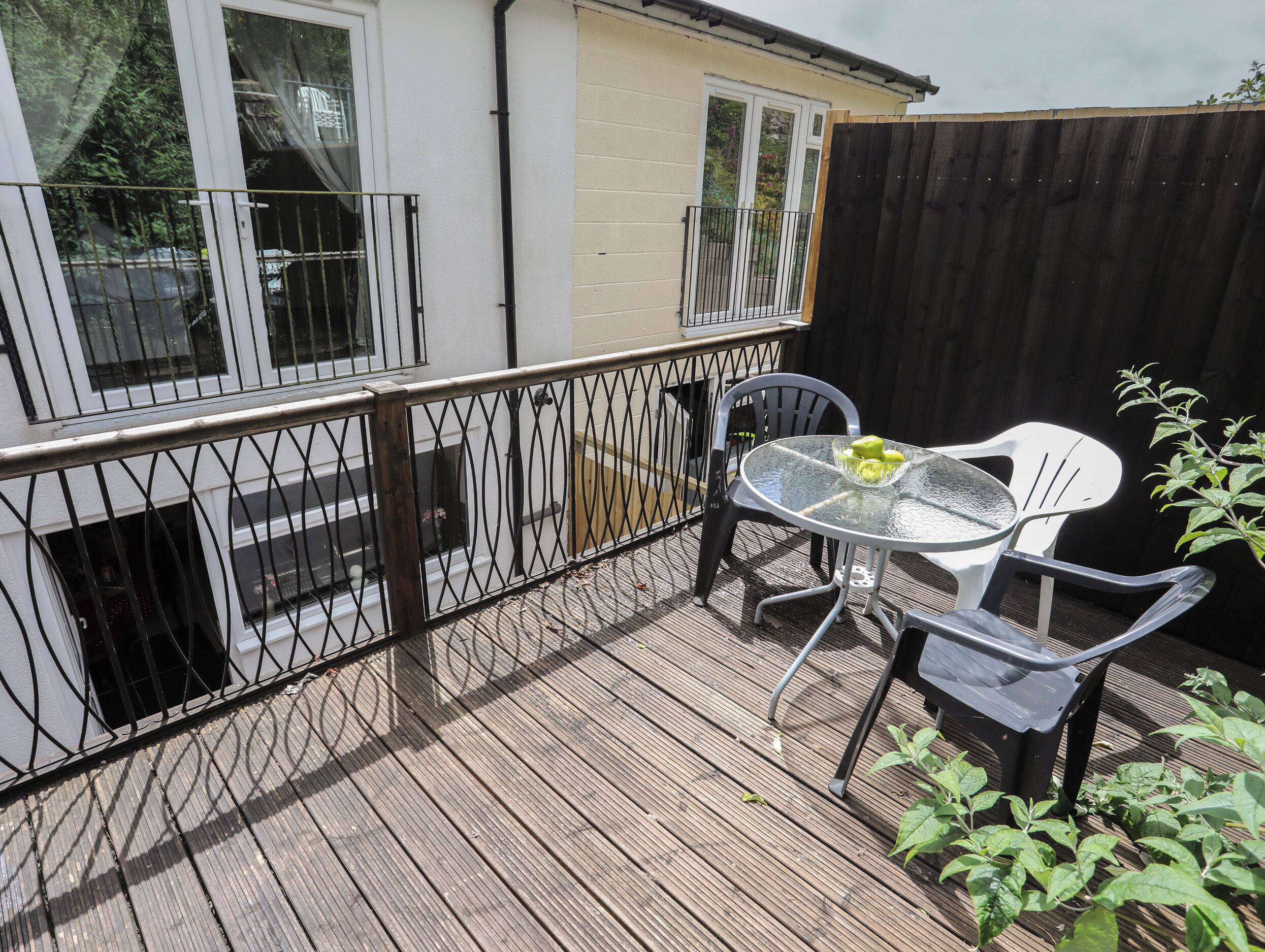 A wooden deck with a glass table and two chairs, a black metal fence, and a wooden fence in the background.