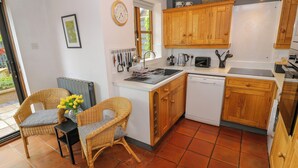 A compact kitchen with wooden cabinets, a dishwasher, and a dining area with wicker chairs. - Old Town Cottage (Stratford-upon-Avon)