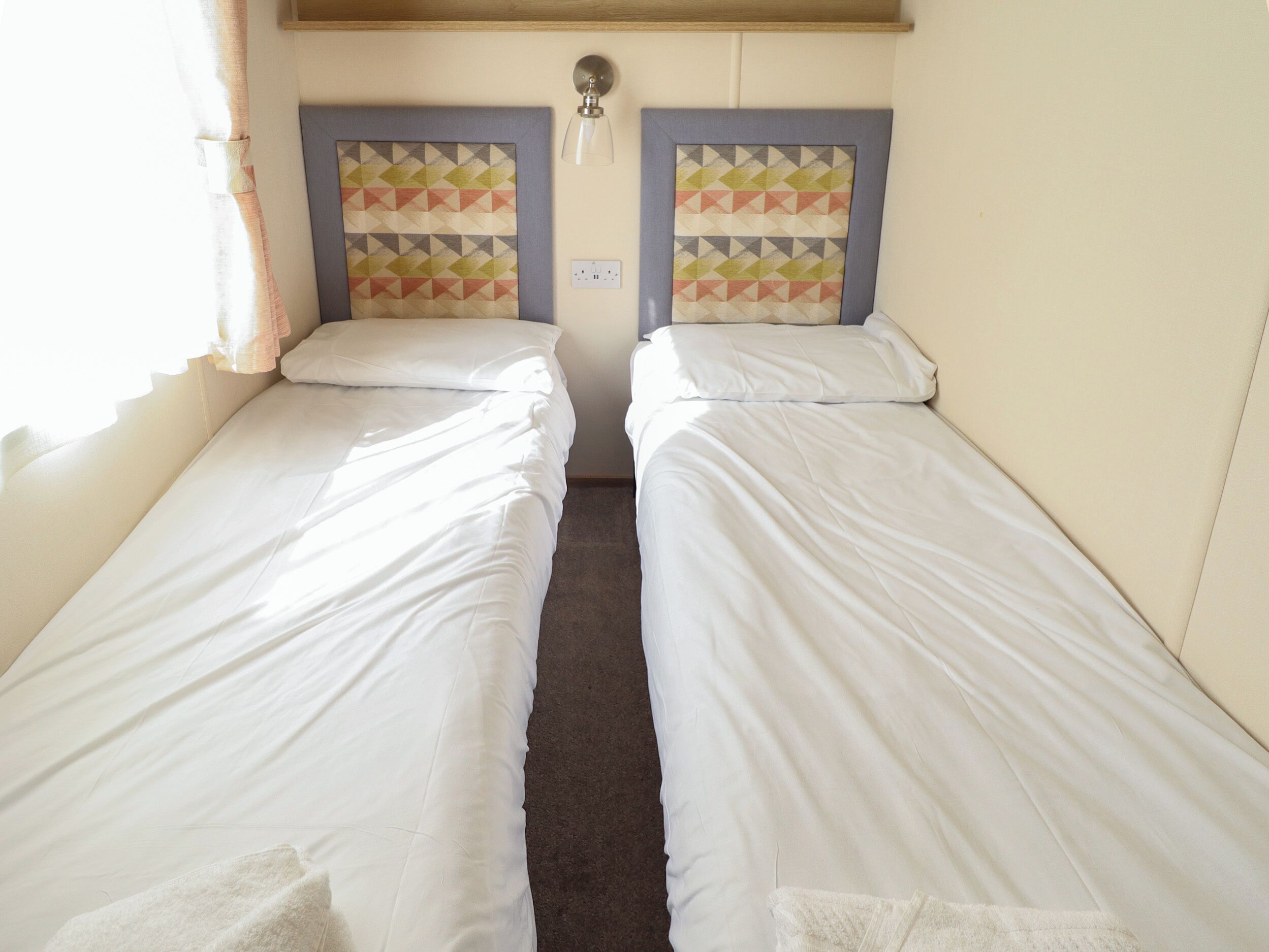 Une chambre avec deux lits simples, chacun étant équipé d’un oreiller et d’une couverture. Un luminaire est installé au-dessus des lits et une prise de courant se trouve sur le mur.