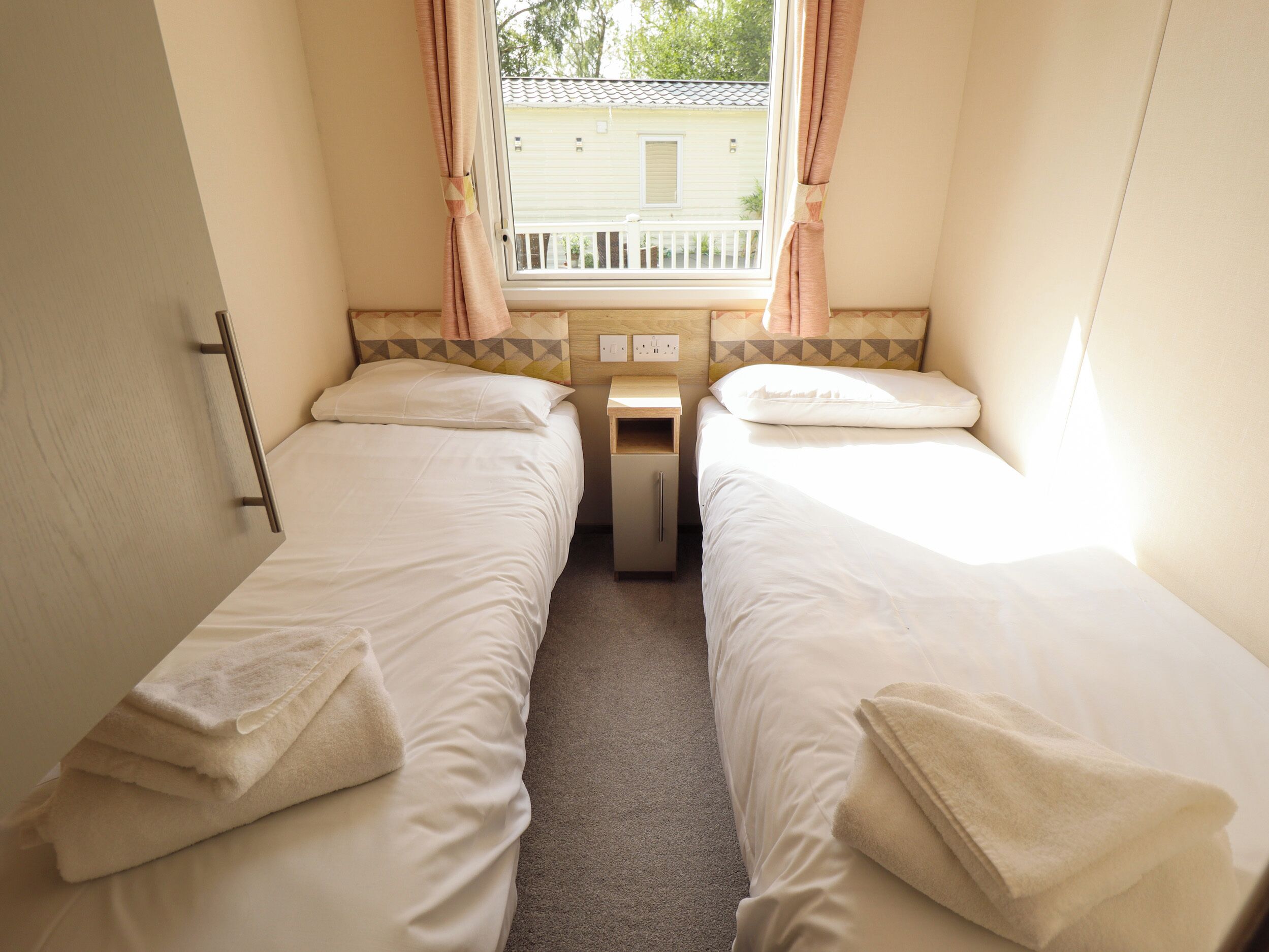 A small, clean room with two single beds, a nightstand, a window with curtains, and a view of a house outside.