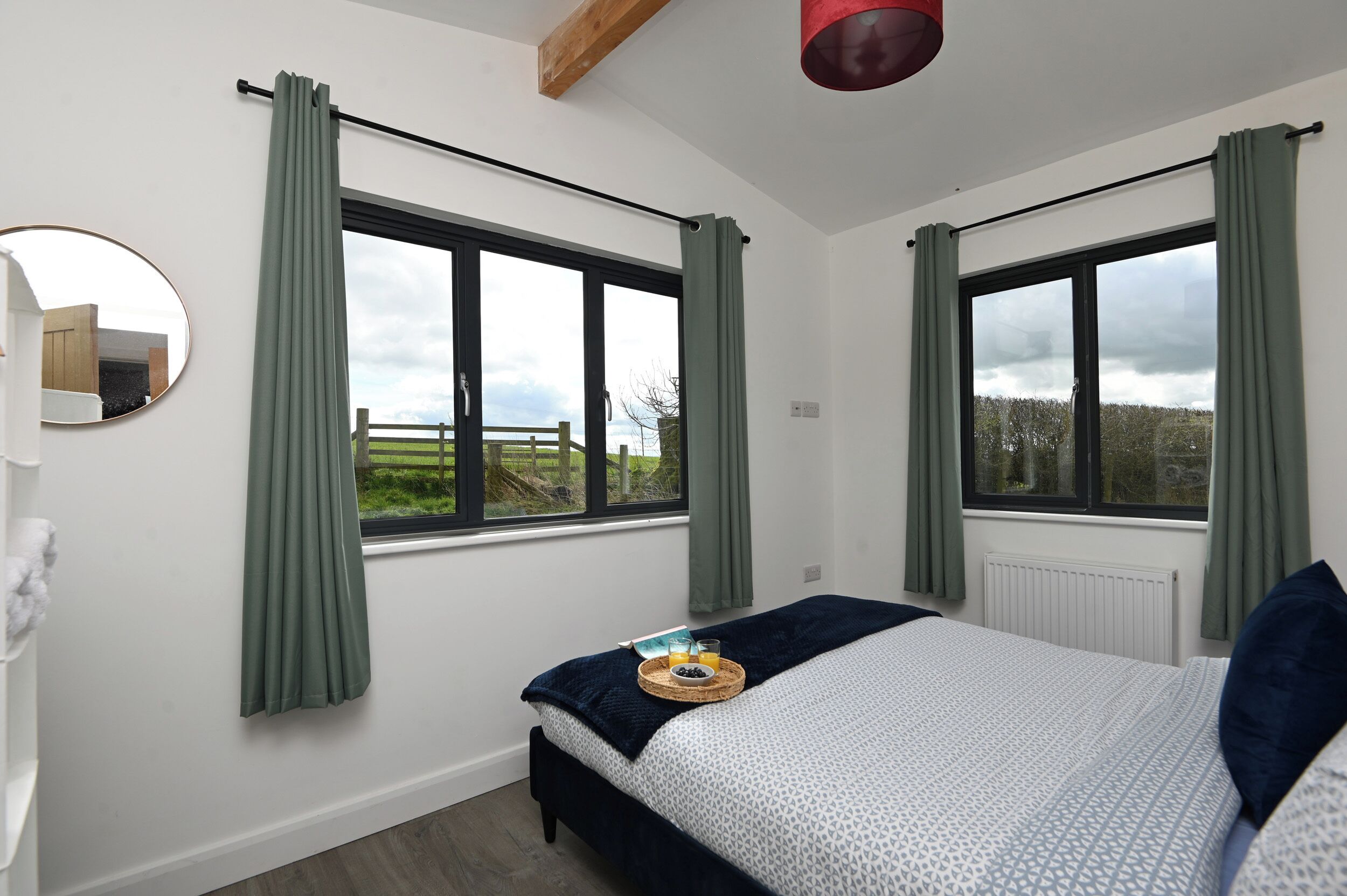 Une chambre avec deux fenêtres, un lit recouvert d’une couverture bleue, une table de chevet avec un plateau contenant un verre de jus d’orange et un livre, et un miroir accroché au mur.