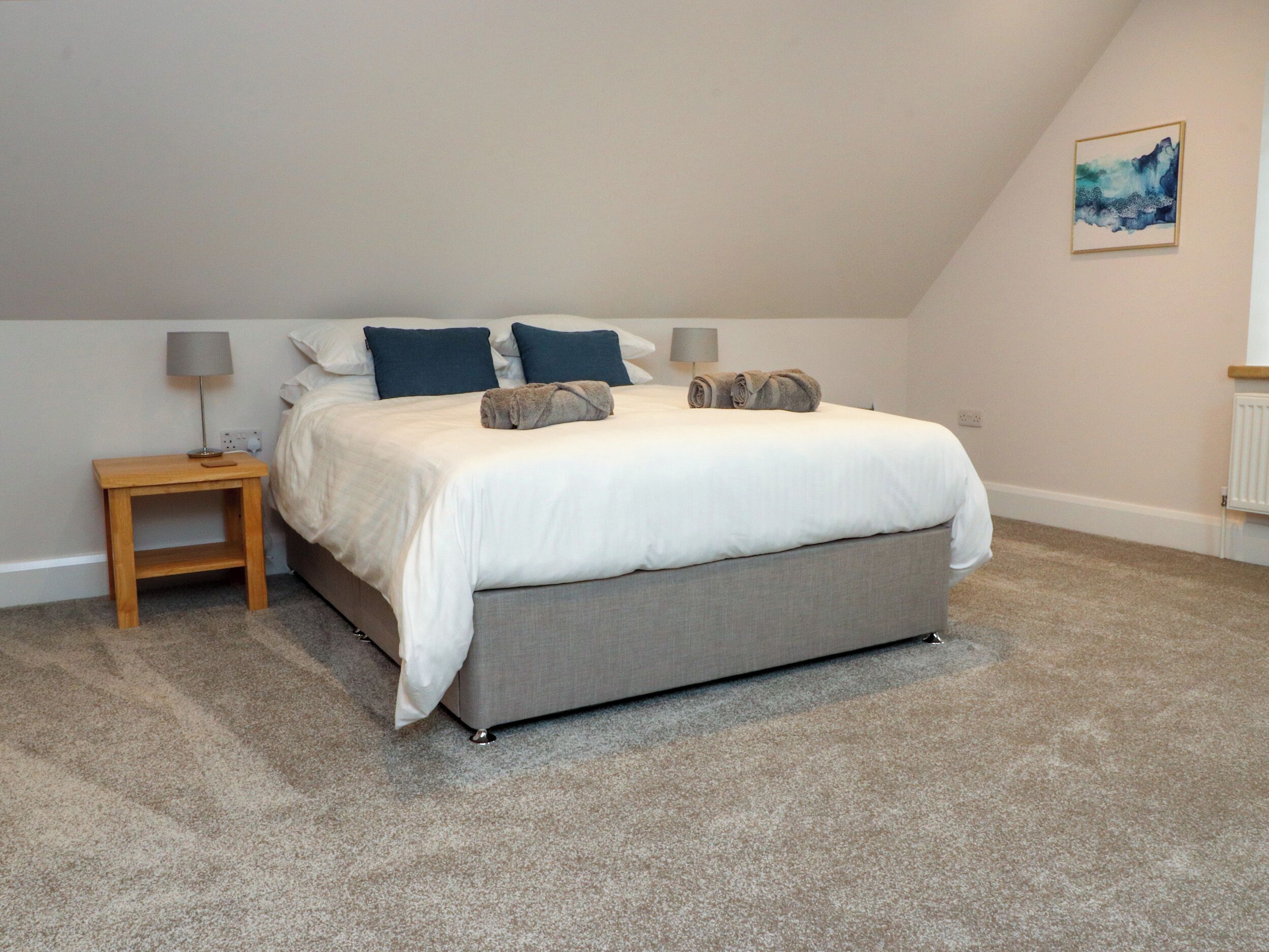 Quarto com cama, mesa de cabeceira, abajur e um quadro na parede.