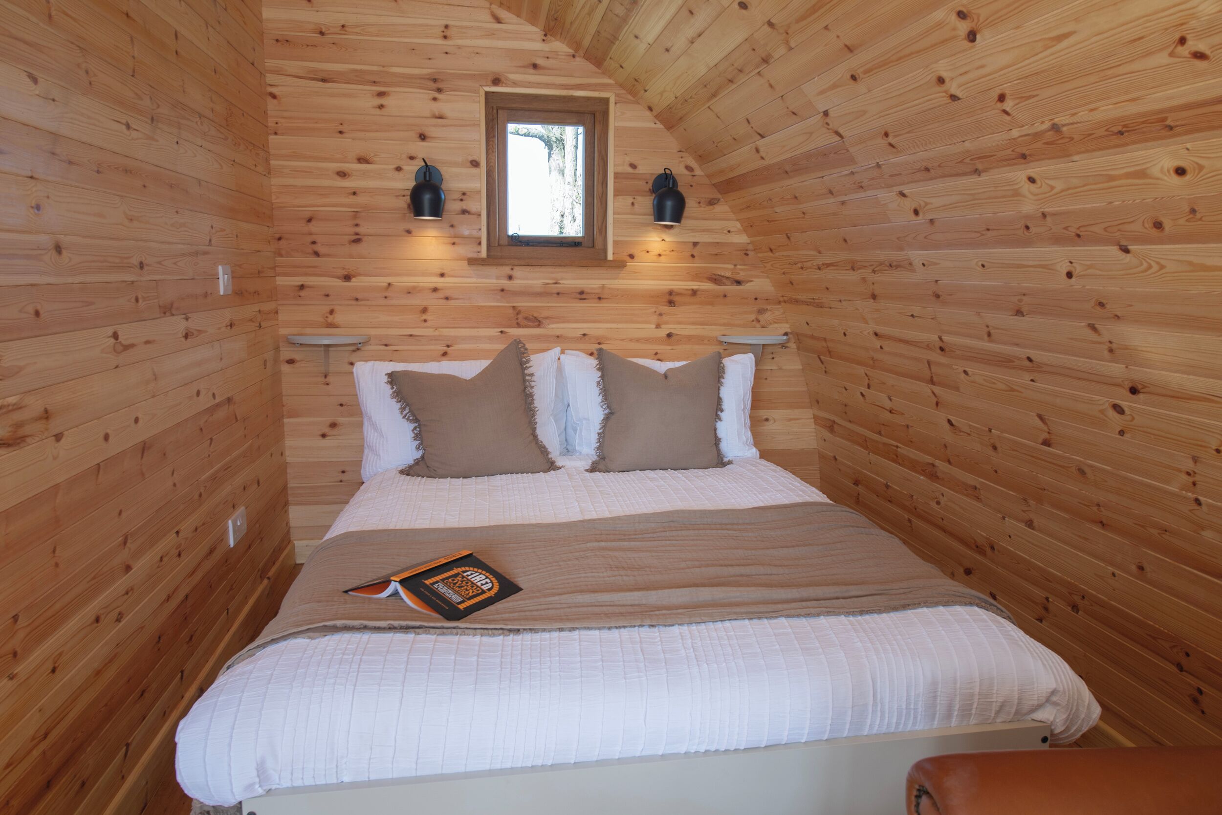 A wooden cabin bedroom with a bed, pillows, a window, and wall-mounted lights.