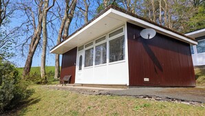 Ein kleines, eingeschossiges Gebäude mit weißer Tür und Fenstern, umgeben von Bäumen und einer Rasenfläche.