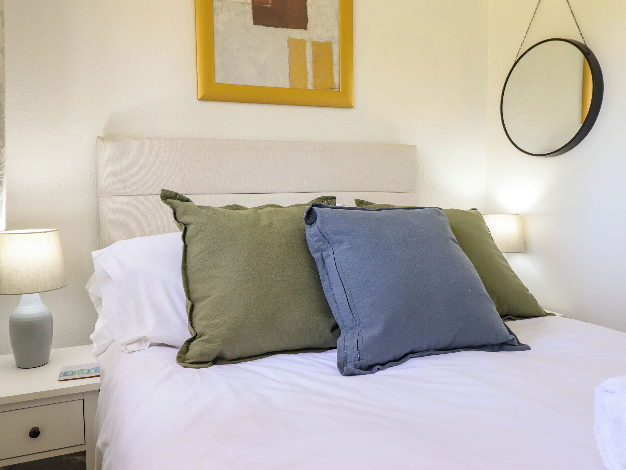A neatly made bed with white linens, two pillows in green and blue, a bedside table with a lamp, and a wall-mounted mirror.