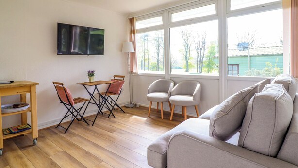 Ein Wohnzimmer mit einer Couch, einem an der Wand montierten Fernseher, einem Esstisch mit Stühlen und großen Fenstern mit Blick auf Bäume und ein Gebäude.
