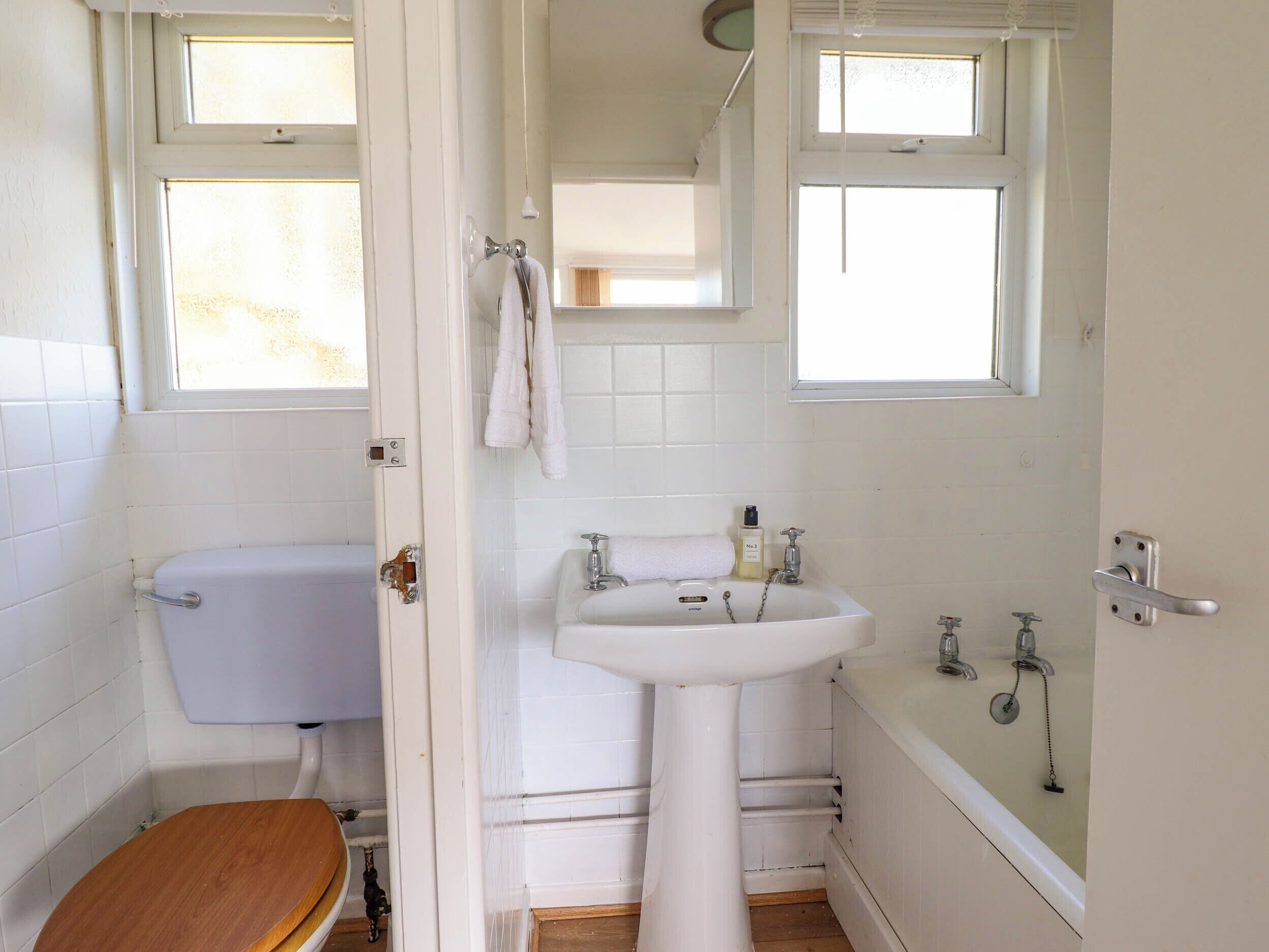 A small, white bathroom with a toilet, sink, and bathtub. There are windows above the sink and bathtub, and a towel hanging on the wall.
