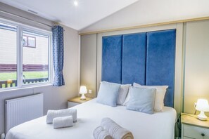 A bedroom with a large bed, two bedside tables with lamps, a window with curtains, and a view of a garden. - Lodge 55 at Riviera Bay Coastal Retreat (Brixham)