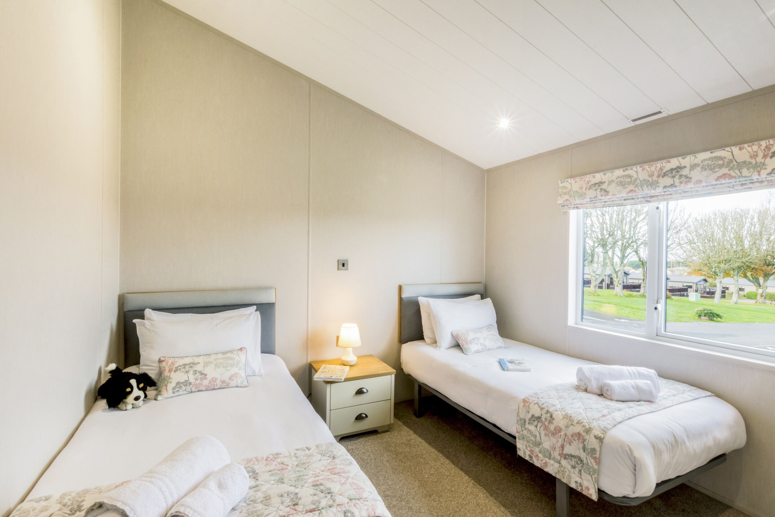 Habitación de hotel con dos camas, una mesita de noche con lámpara y una ventana con vista a árboles.