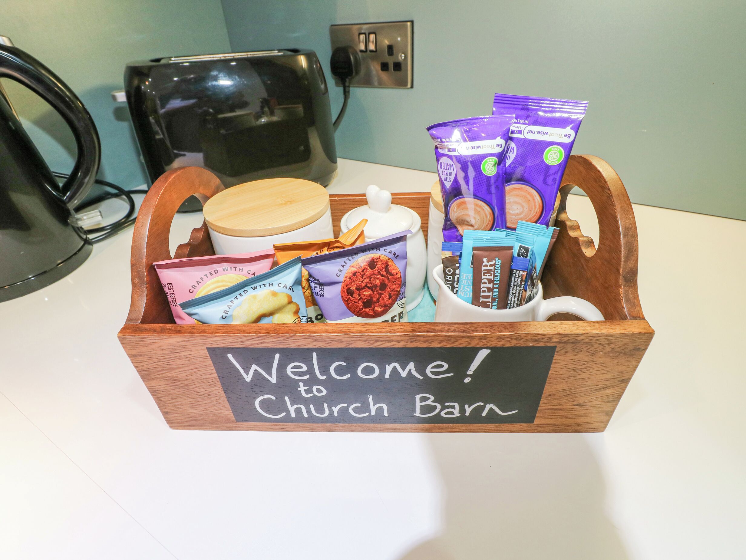 Una cesta de madera con un letrero que dice 'Welcome to Church Barn' que contiene diversas golosinas empaquetadas y una taza blanca.