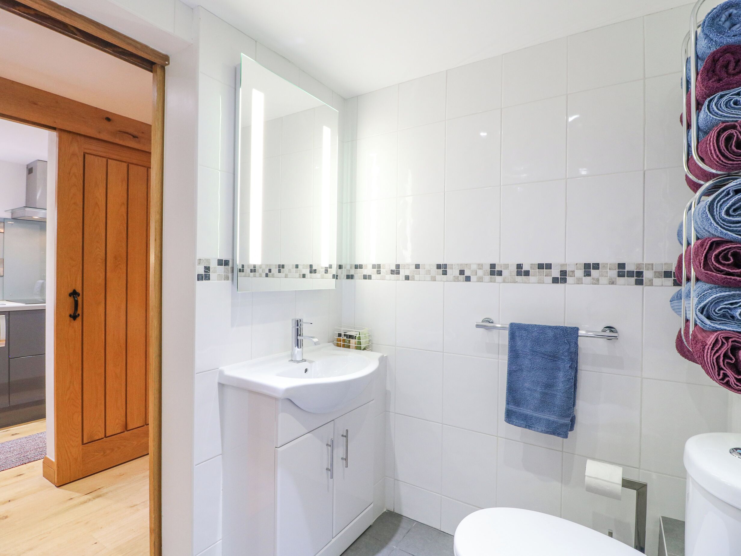 Een moderne badkamer met een wit wastafelmeubel, spiegel en toilet. Een houten deur leidt naar een andere kamer.