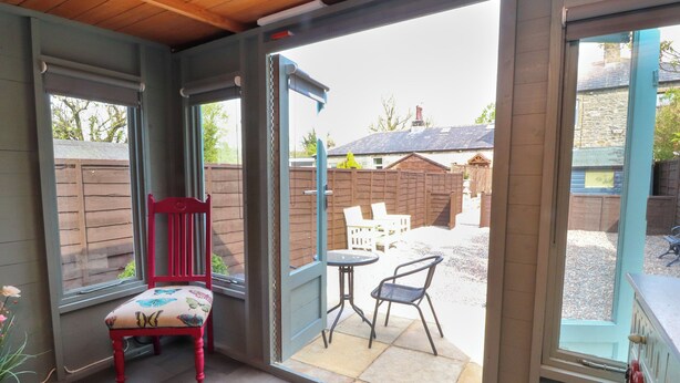 Una stanza con soffitto in legno, una sedia rossa, un tavolo e una porta scorrevole in vetro che dà su un patio esterno con recinzione e una casa sullo sfondo.