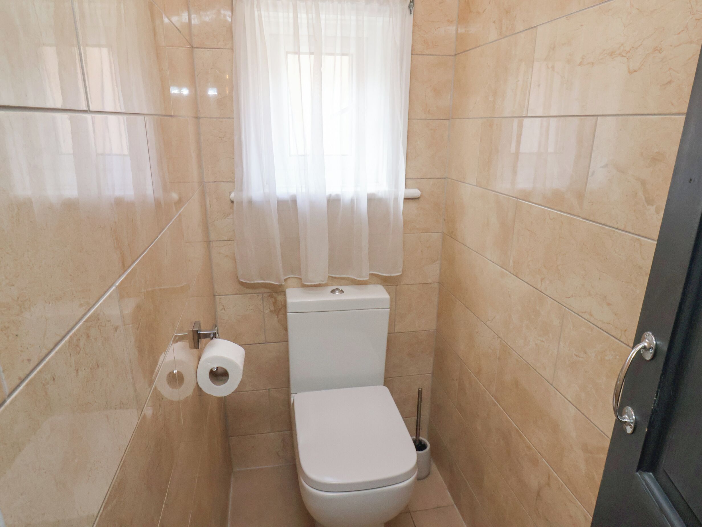 A small, tiled bathroom with a toilet, two rolls of toilet paper, a toilet brush, and a white curtain.
