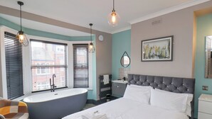 Une chambre à coucher moderne avec un grand lit, une baignoire séparée, une coiffeuse avec un miroir et un tableau représentant un paysage urbain accroché au mur.