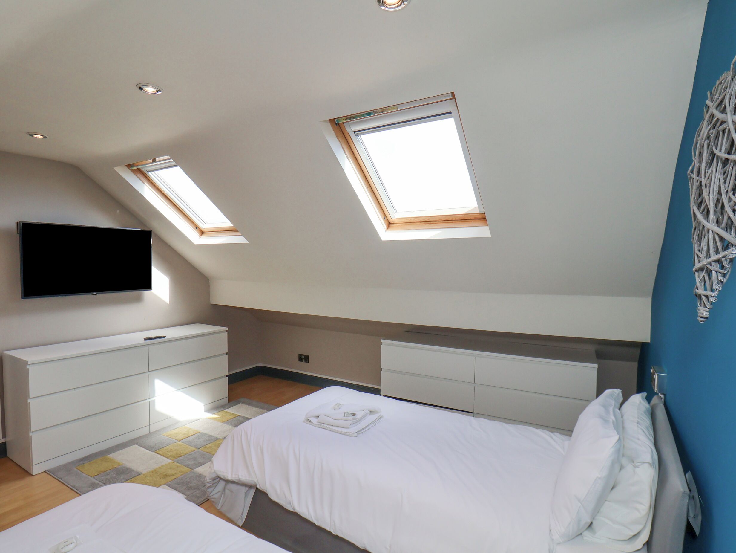Un dormitorio con techo inclinado, tragaluces, televisor de pantalla plana, cómoda blanca y cama con ropa de cama blanca.