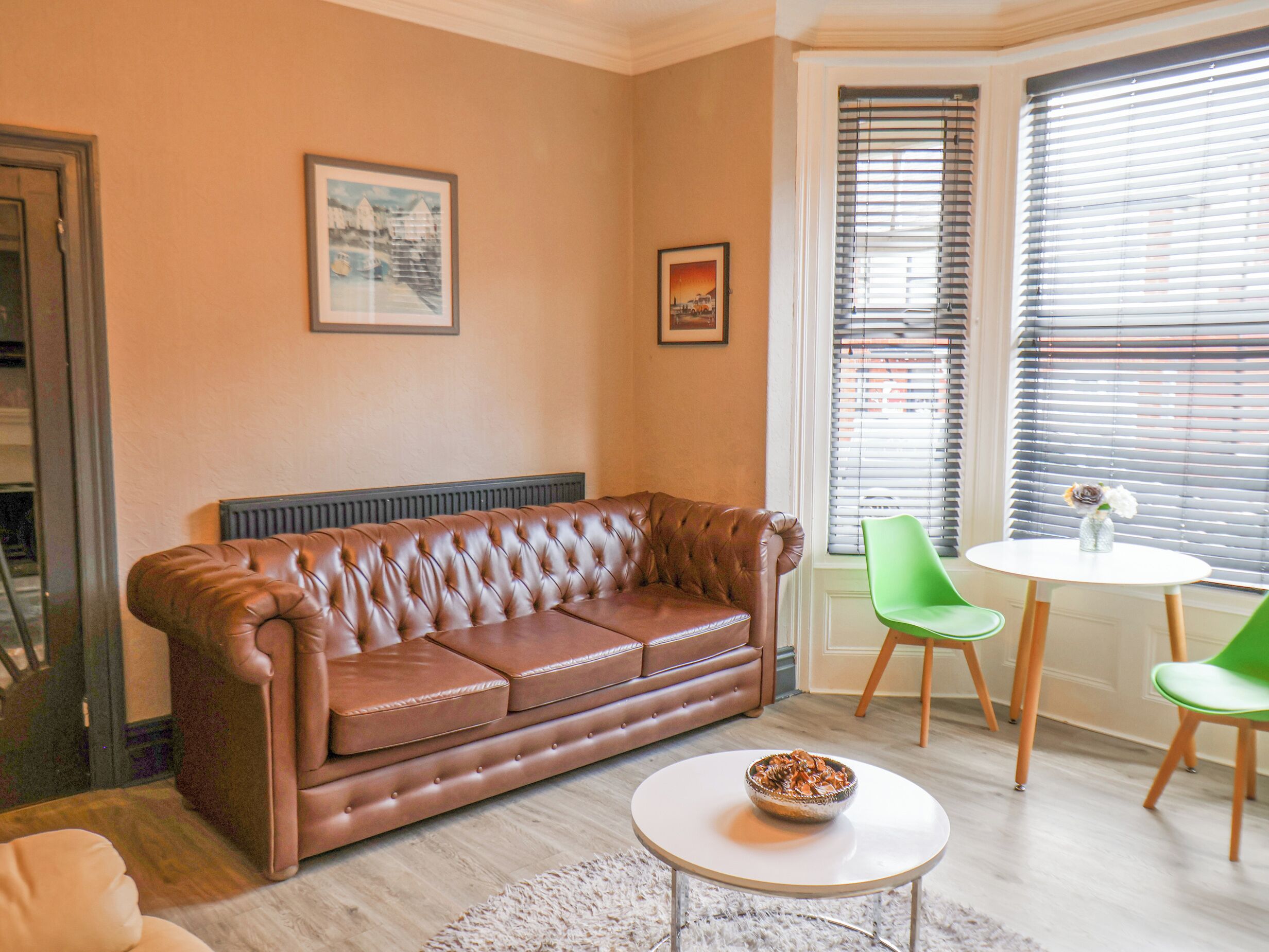 A living room with a brown leather sofa, a small white table with a bowl, green chairs, and a window with blinds.