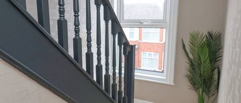 Un escalier avec des balustrades noires menant à un palier, une plante en pot sur le palier et une fenêtre donnant sur un bâtiment à l’extérieur.